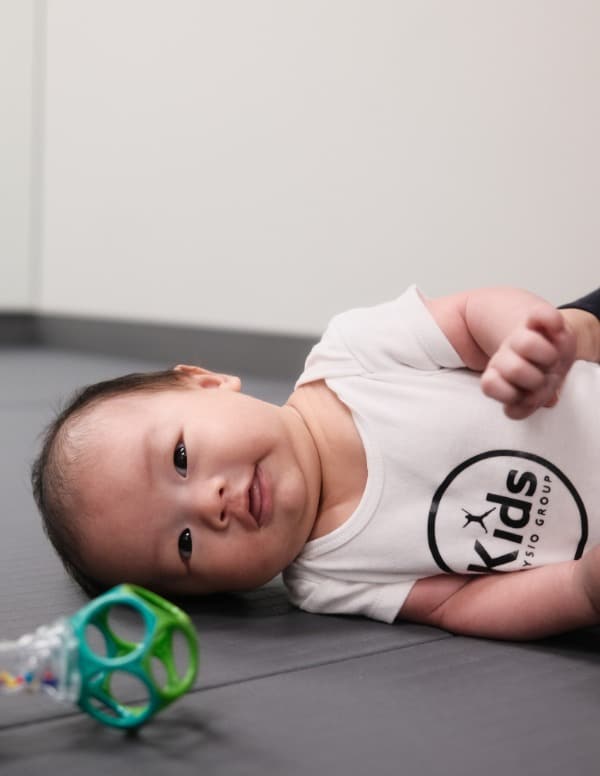 A baby wearing a white onesie with the Kids Physio Group logo, lying on their side facing the camera. They are smiling.