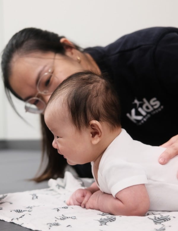 An infant on its tummy being treated by a physiotherapist who is on their side for Torticollis