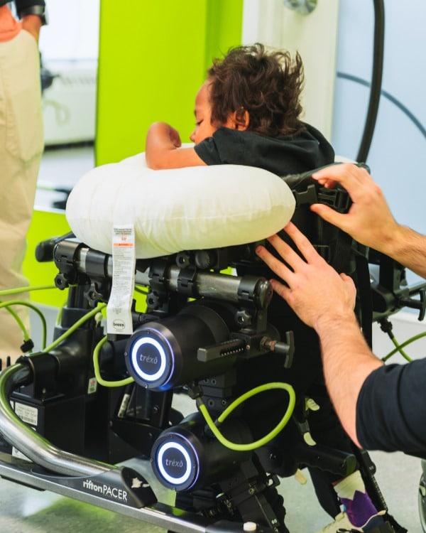 A child being securely fitted into a Trexo robotic gait trainer by a pediatric physiotherapist to practice walking and upright movement.
