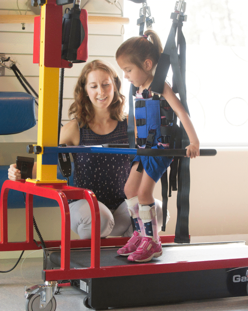 A young girl wearing leg braces using a supported treadmill with a harness during a pediatric physiotherapy session for cerebral palsy in infants or children.