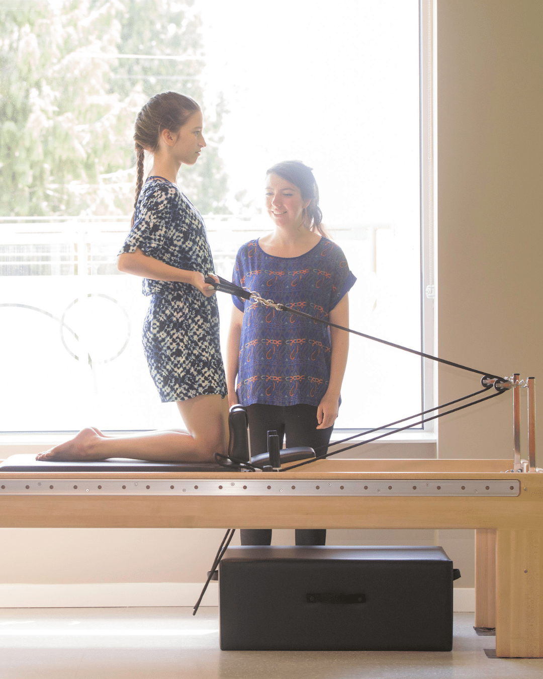 A young girl kneeling on a clinical reformer while performing resistance exercises under the supervision of a pediatric physiotherapist.
